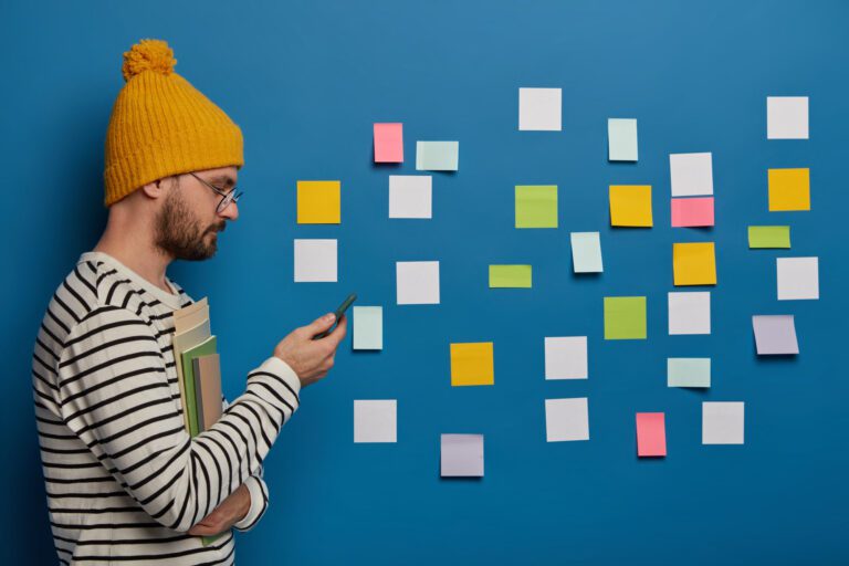 Stylish schoolboy scrolls information in internet via modern cellphone, stands with books indoor, turns sideways to camera, makes strategy plan, uses sticky notes to remember important material